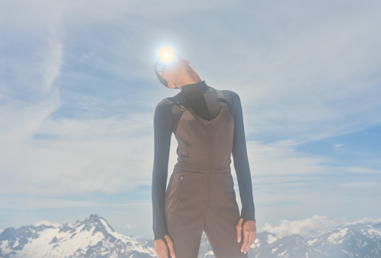 Model in an espresso brown ski jumpsuit with Alpine peaks behind