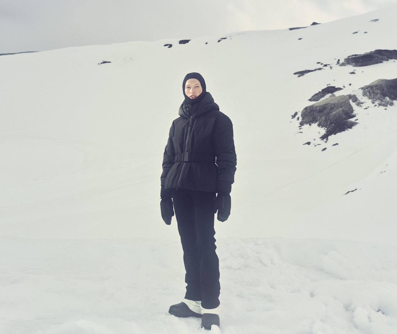 Model in an all-black ski suit and balaclava standing on snow