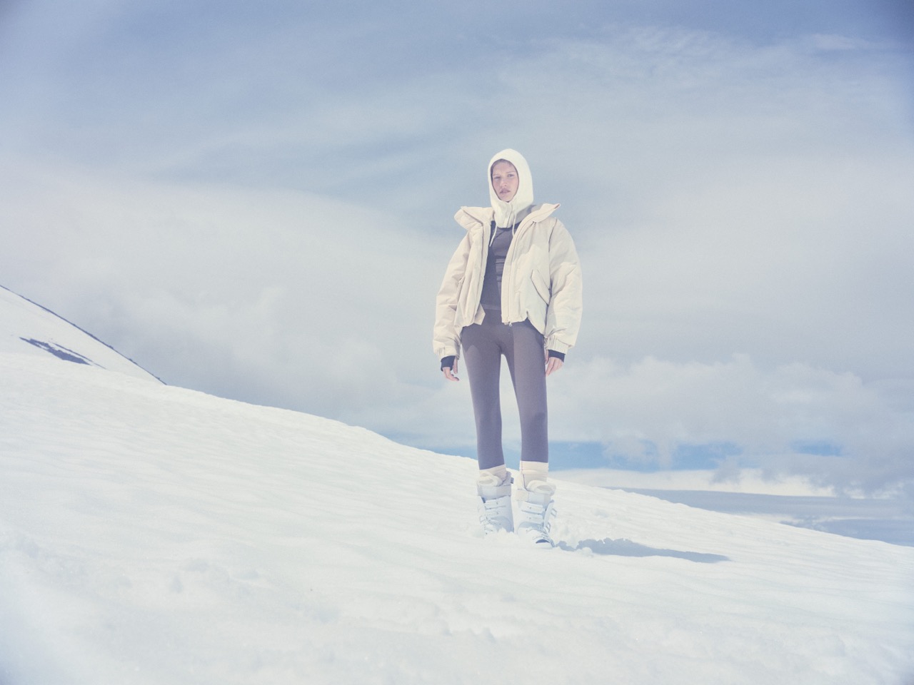 Model in a cream puffer jacket and muted purple ski trousers on a snowy slope