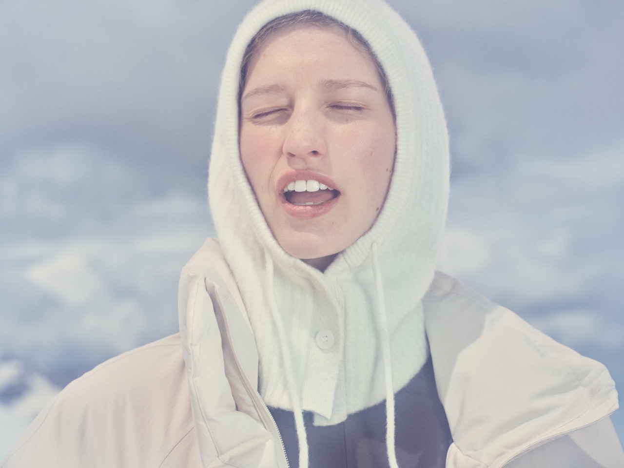 Model in a cream knitted hooded balaclava against a cloudy Alpine sky