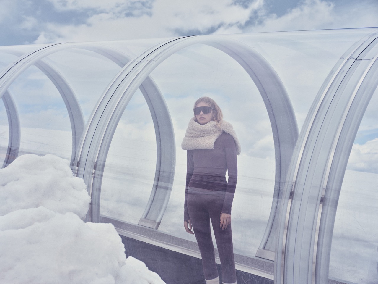 Model in a black ski outfit standing inside a glass tunnel above the clouds